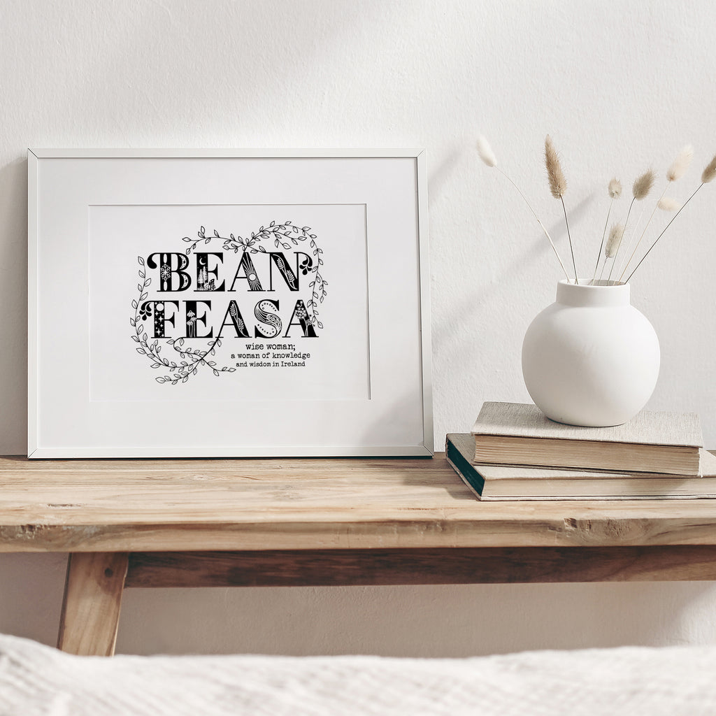 bean feasa screen print mounted in a white frame leaning against the wall and sitting on a  wood bench beside a white case with dried flowers