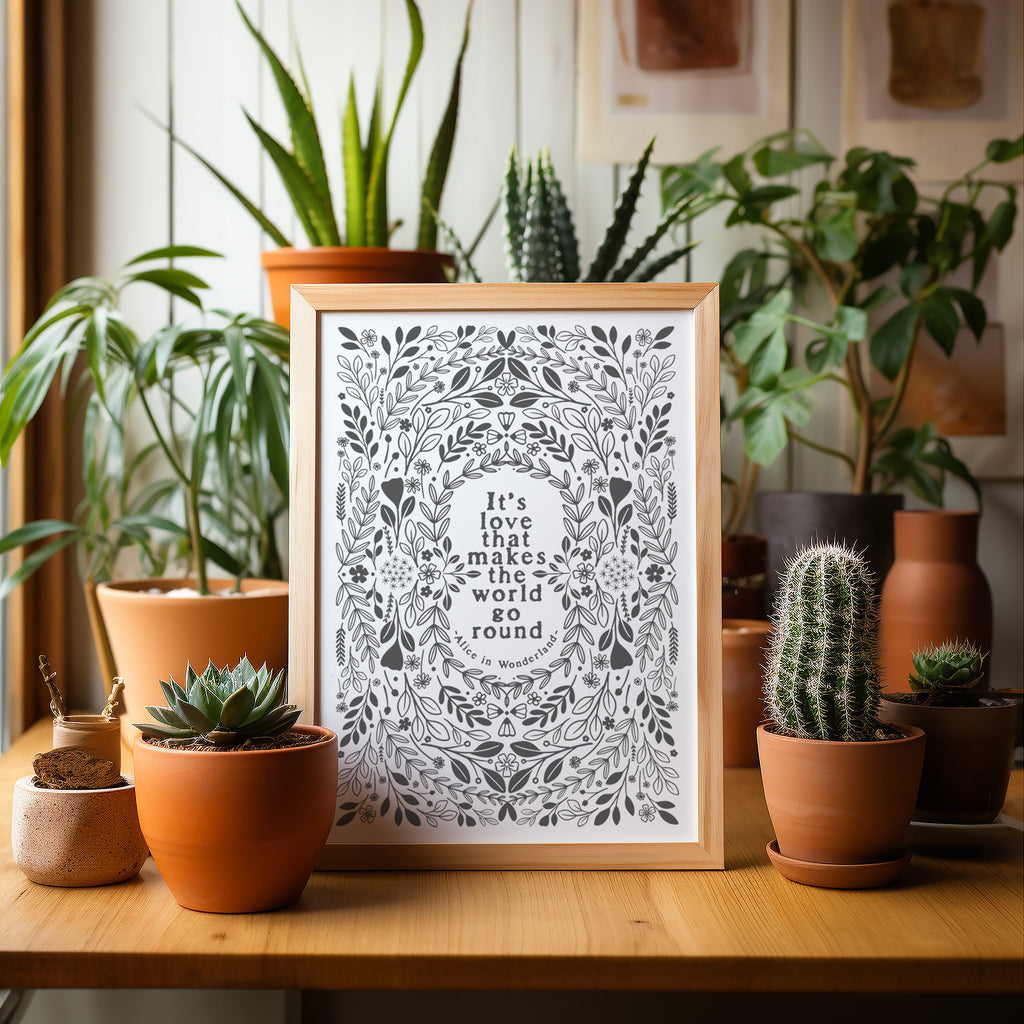 Grey and white screen print in a wood frame sitting on  a table surrounded by potted plats and cacti