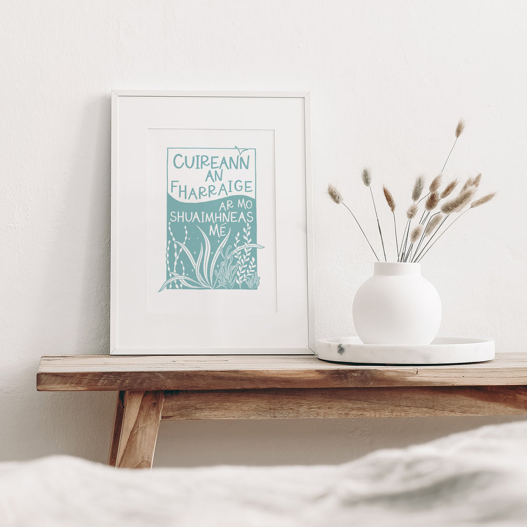 Blue and white Irish screen print of the sea and coral mounted in a  white frame. It is leaning against a white wall and sitting on a wood bench beside a white vase with dried flowers