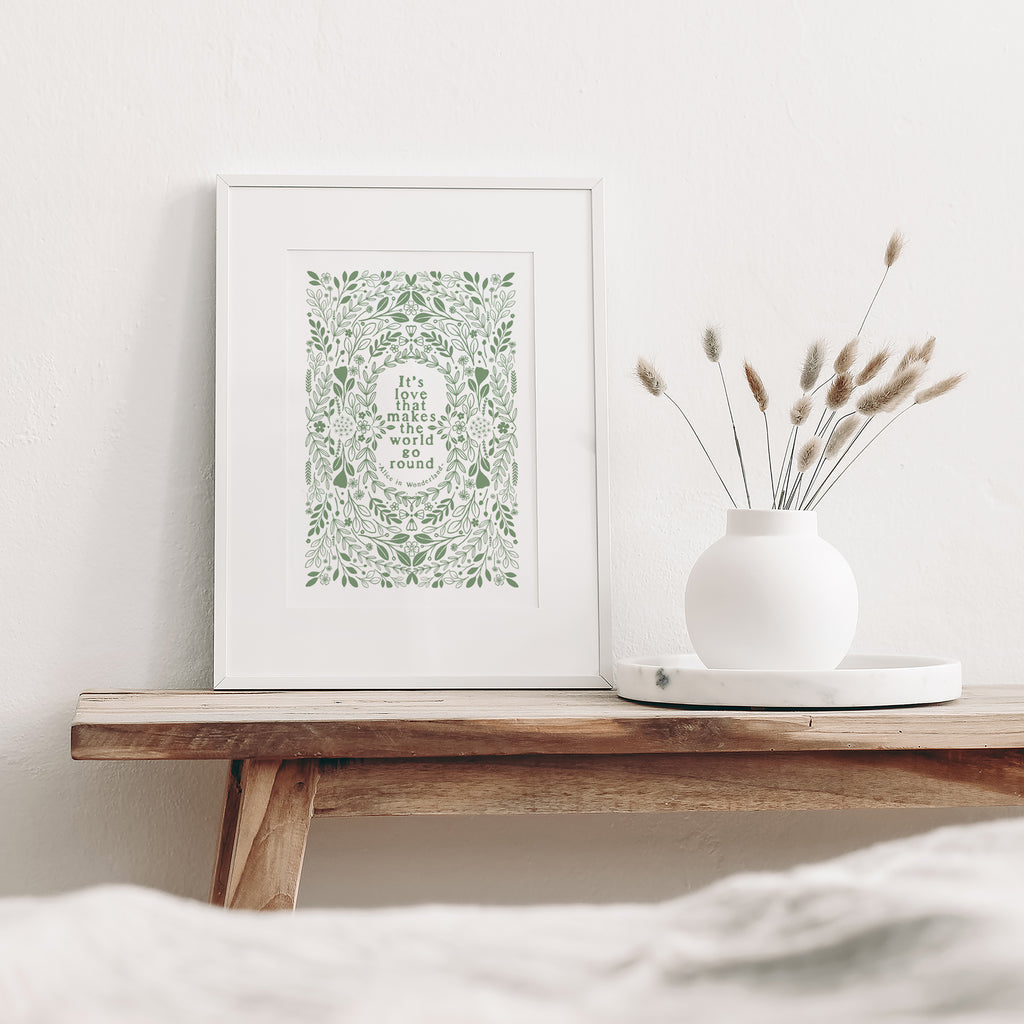 Green and white screen print mounted in a white frame leaning on a white wall. It is sitting a wood bench beside a white vase with dried flowers