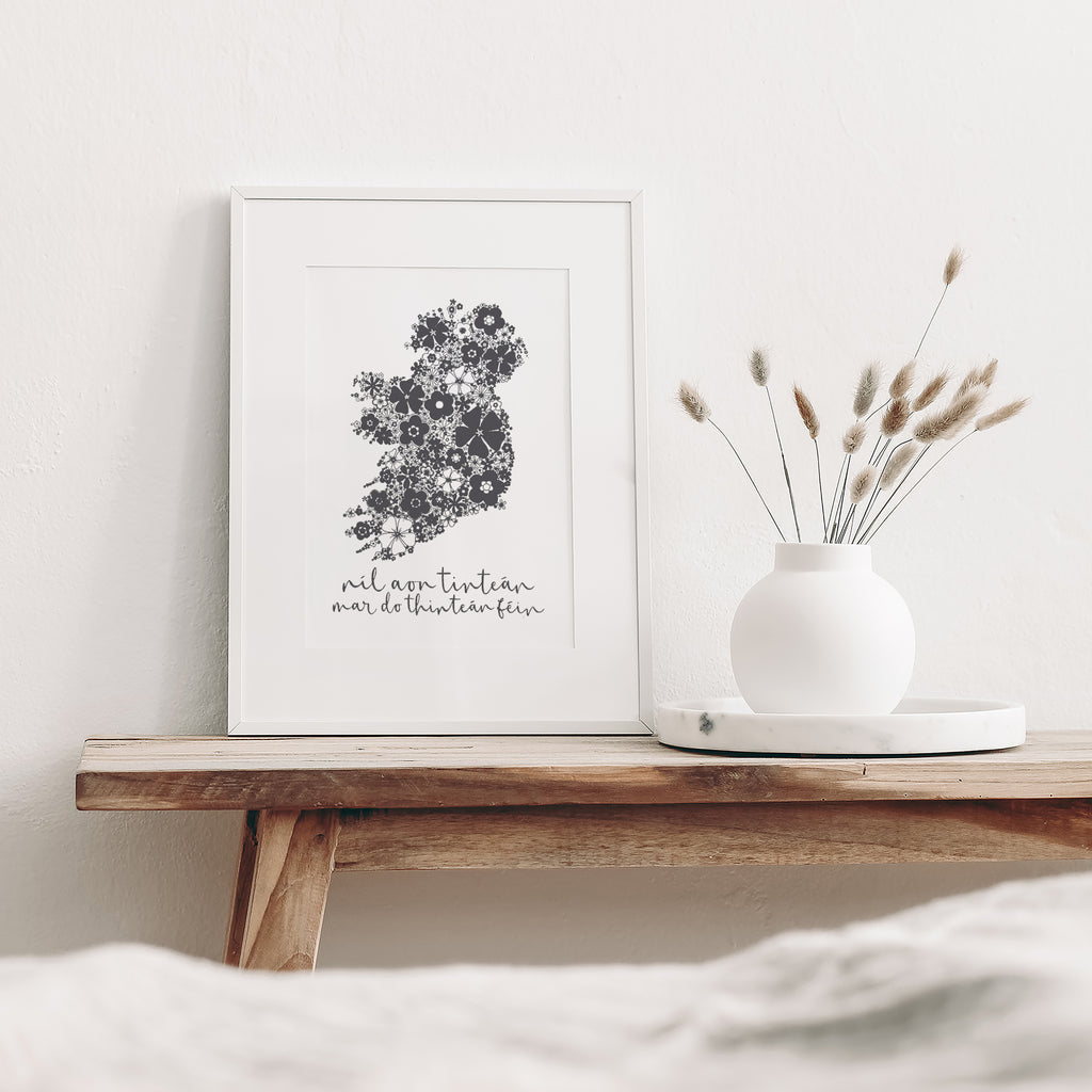 Grey Irish screen print mounted in a white frame leaning against a white wall. It is sitting on a wood bench beside a white vase with dried flowers
