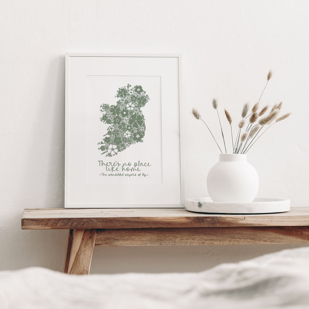 Green Ireland screen print mounted in a white frame leaning against a white wall. It is sitting on a wood bench beside a white vase with dried flowers
