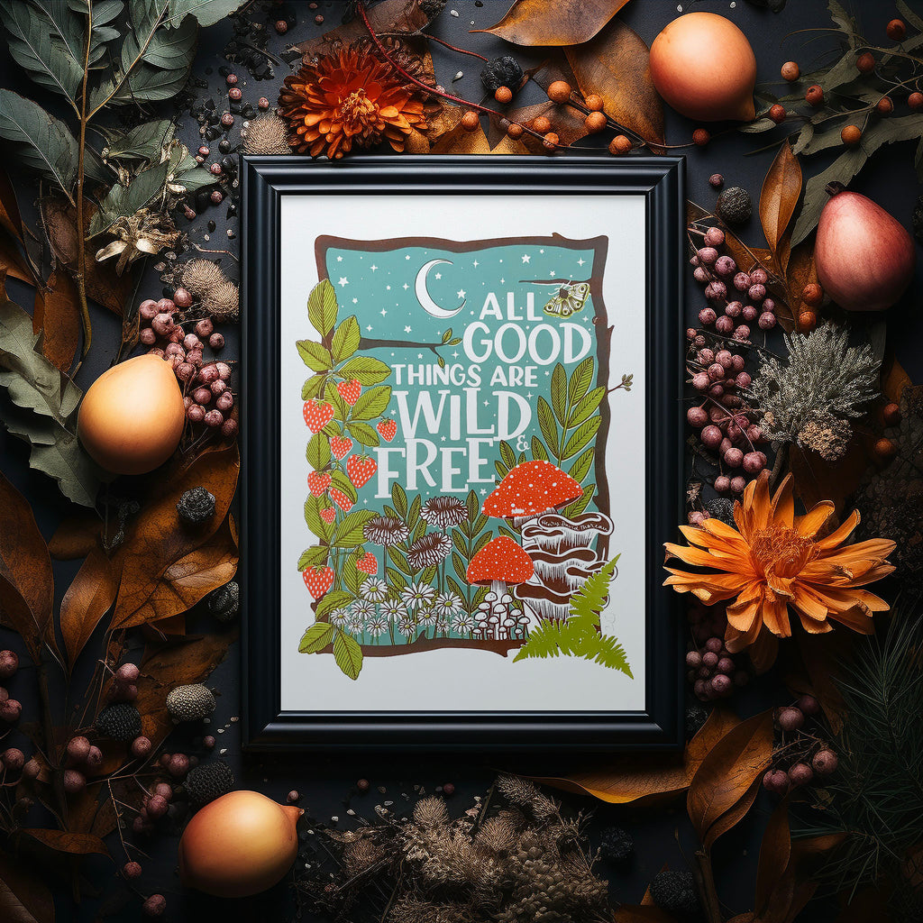 Wild and free screen print in a black frame sitting on a dark table surrounded bye autumnal flowers and foliage