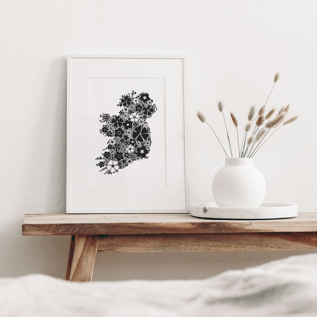Black floral Ireland screen print mounted in a white frame leaning against a white wall. It is sitting on a wood bench beside a white vase with dried flowers