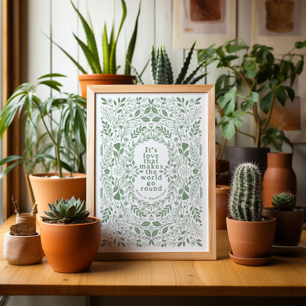 Green and white screen print in a wood frame sitting on a table and surrounded by potted plants and cacti