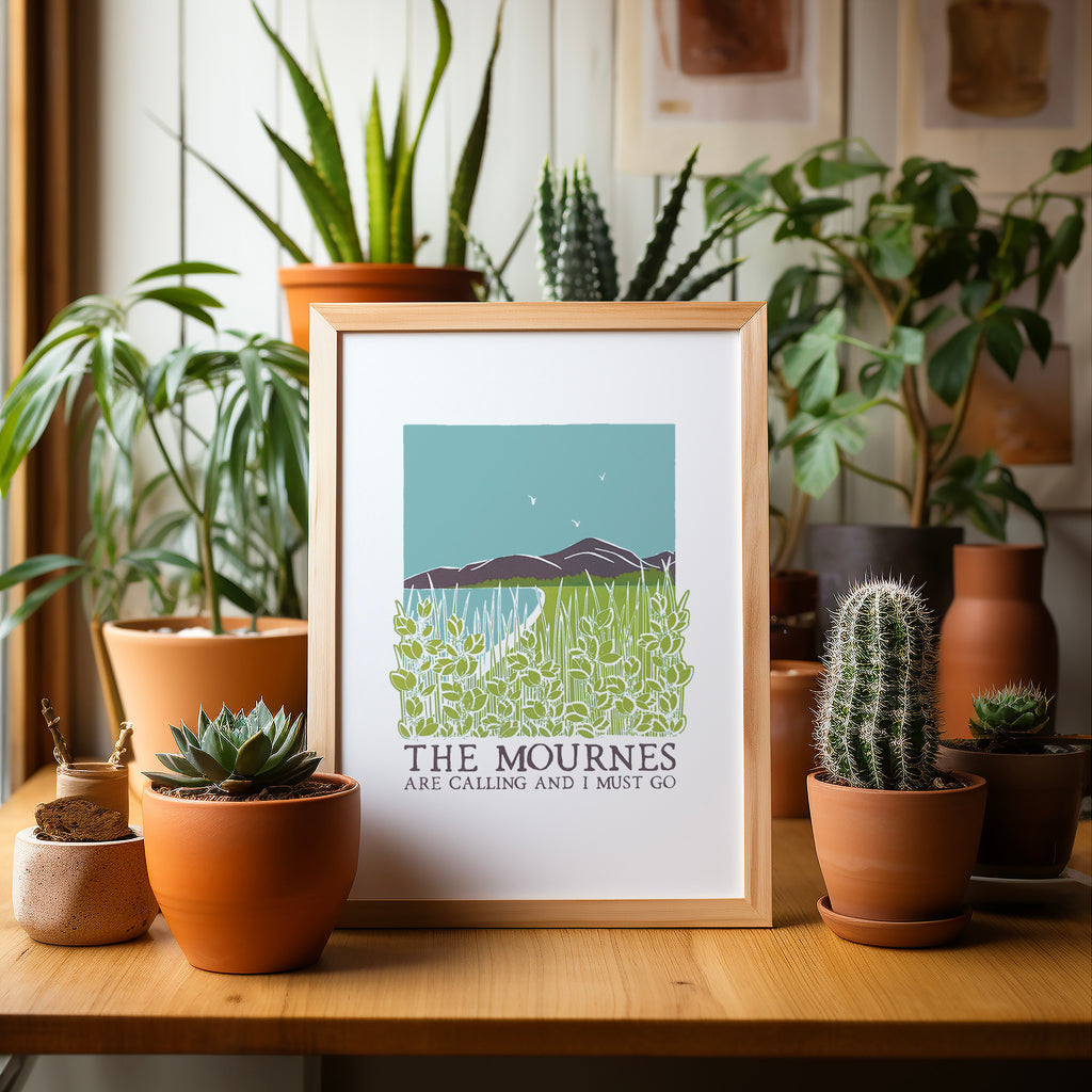 The Mournes are calling screen print in a wood frame sitting on a wood table surrounded by potted plants and cacti
