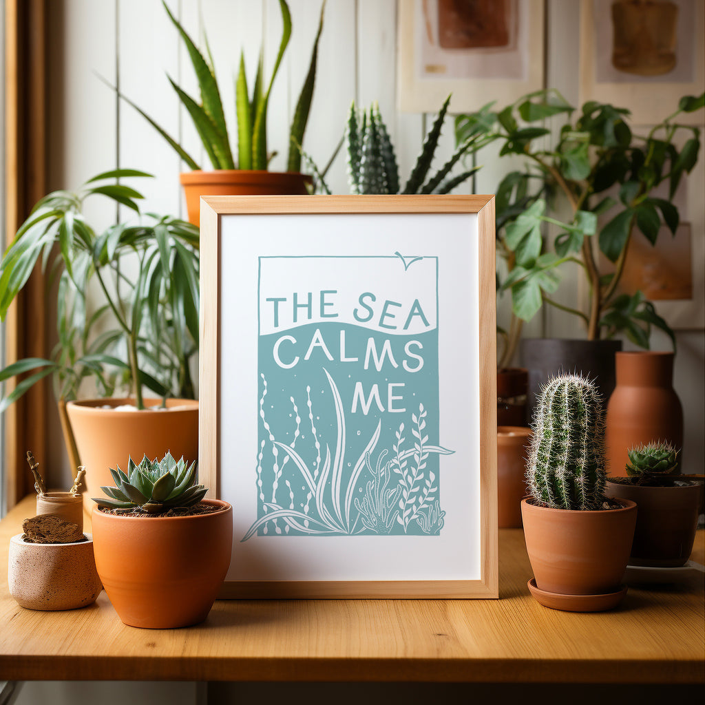 Blue and White Sea screen print in a wood frame sitting on a wood table surrounded by potted plants and cacti