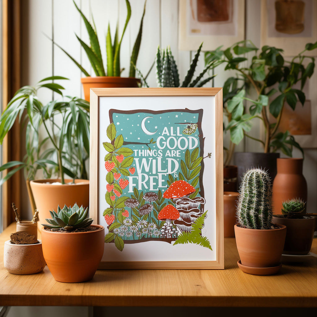 Our 'All good things are wild and free' screen print in an A4 frame, sitting on a table surrounded by potted cacti and plants