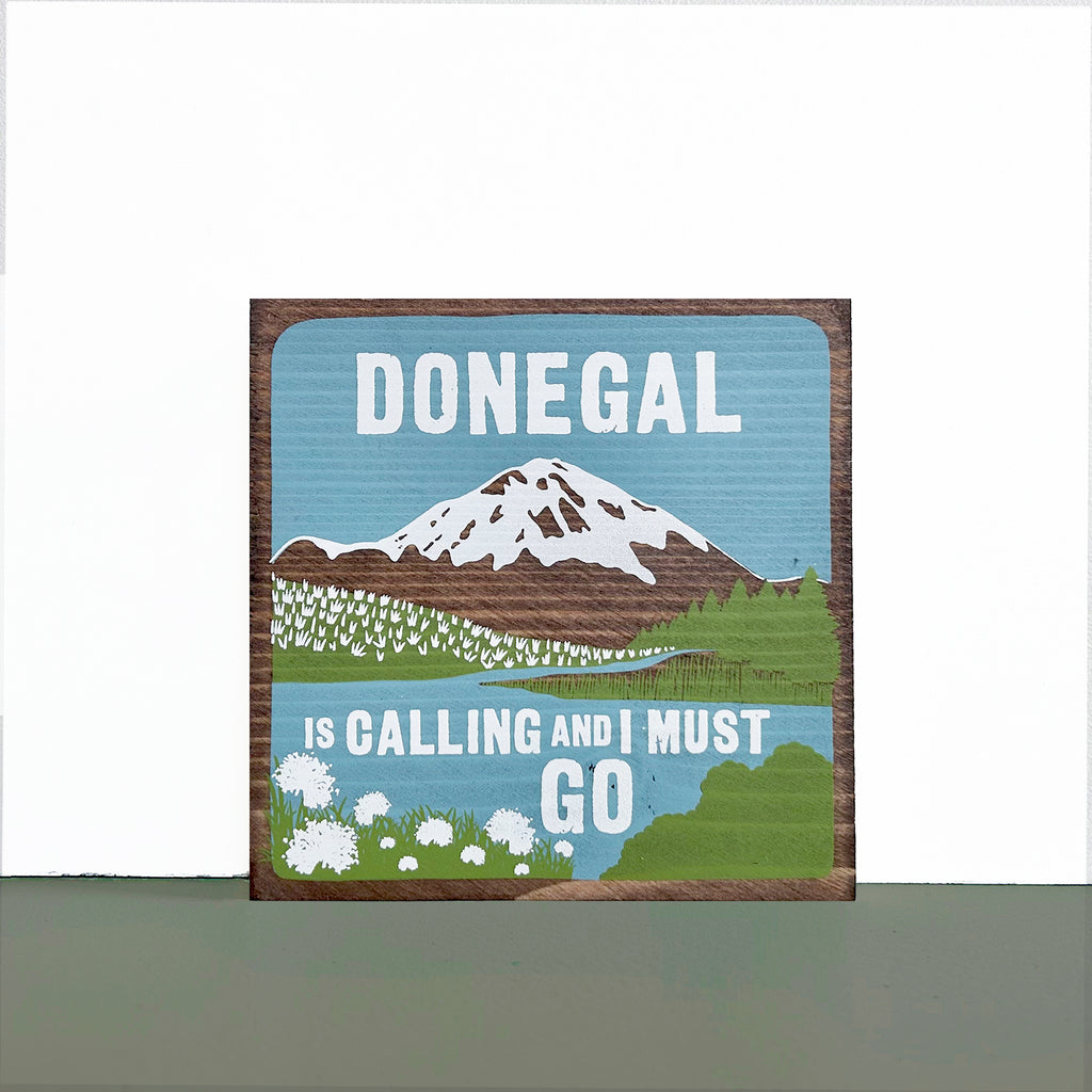 Square wood sign, with Donegal design, sitting on a green shelf against a white wall