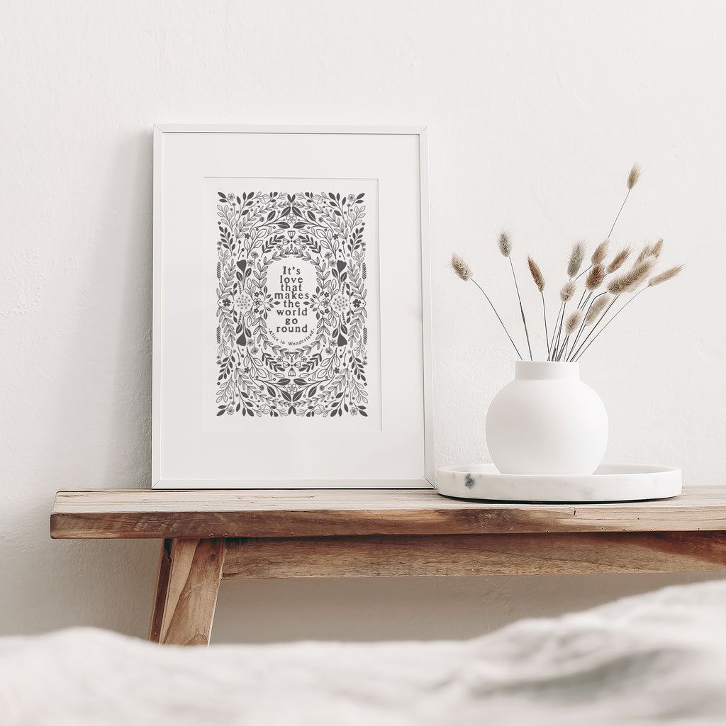 Grey and white print mounted in a white frame leaning against a white wall. It is sitting on a wood bench beside a white vase with dried flowers