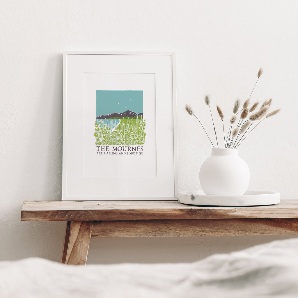 The Mournes are calling screen print mounted in a white frame leaning against a white wall. It is sitting on a wood bench beside a white vase with dried flowers