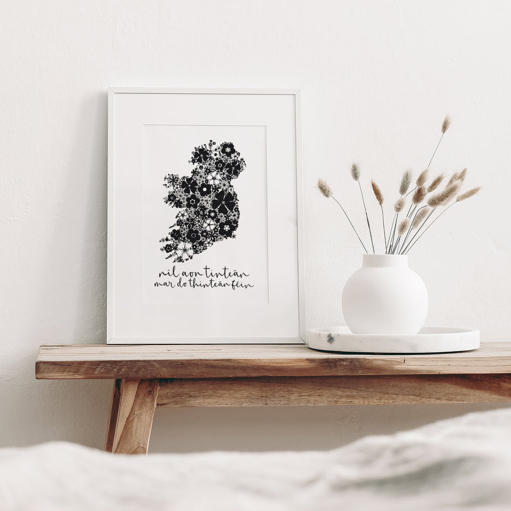 Black Irish screen print mounted in a white frame leaning against white wall. It is sitting on a wood bench with a white vase and dried flowers