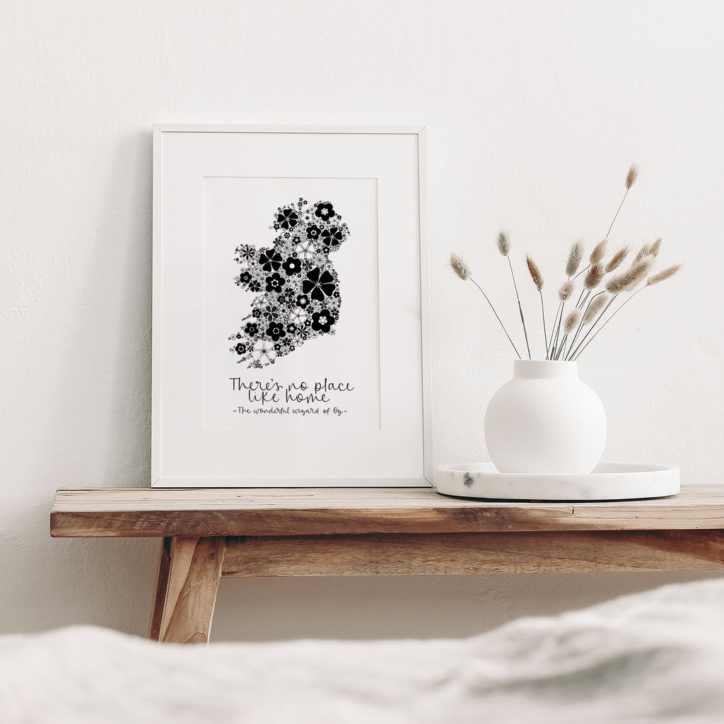 Black Ireland screen print mounted in a white frame leaning against a white wall. It is sitting on a wood bench beside a white vase with dried flowers