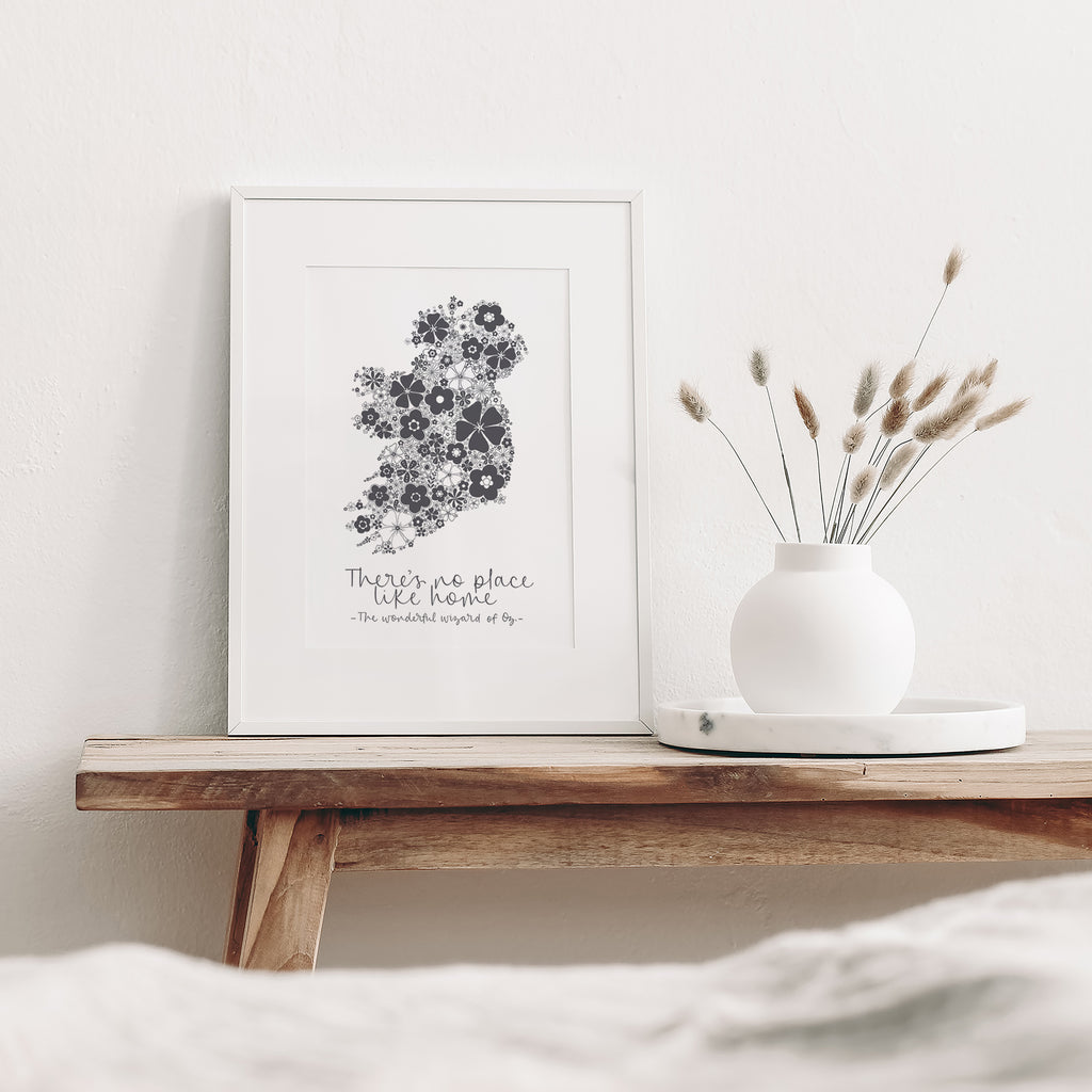 Grey Ireland screen print mounted in a white frame leaning against white wall. It is sitting on a wood bench beside a white vase with dried flowers