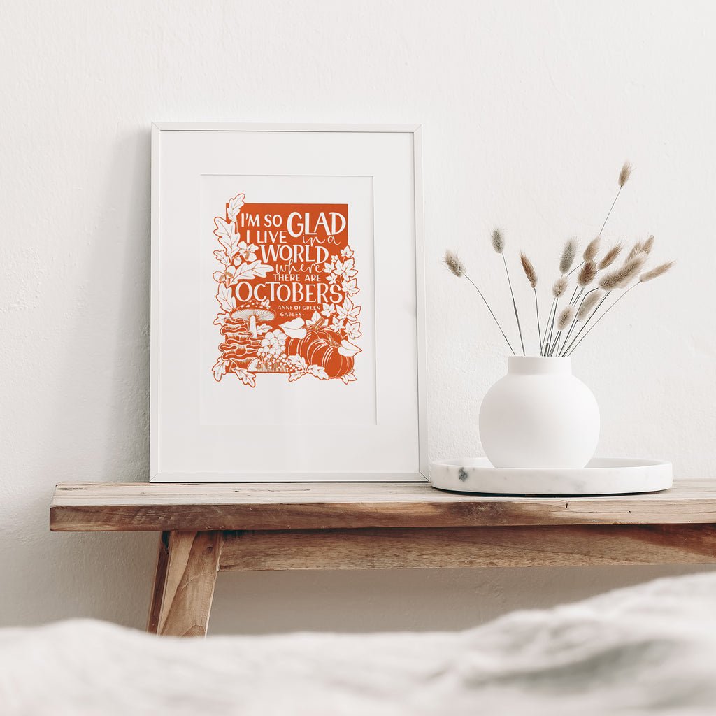 Orange Octobers screen print mounted in a white frame leaning against a white wall. It is sitting on a wood bench beside a white vase with dried flowers