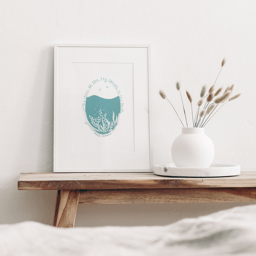 Voice of the sea... screen print mounted in a white frame leaning against a white wall.  It is sitting on a wood bench beside a white vase with dried flowers