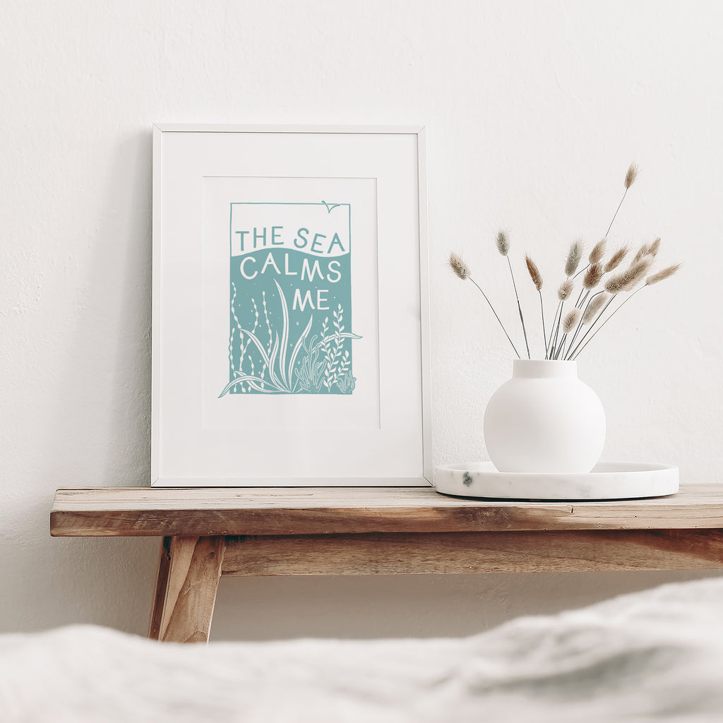 Blue and White Sea screen print mounted in a white frame leaning against a white wall. It is sitting on a wood bench beside a white vase with dried flowers