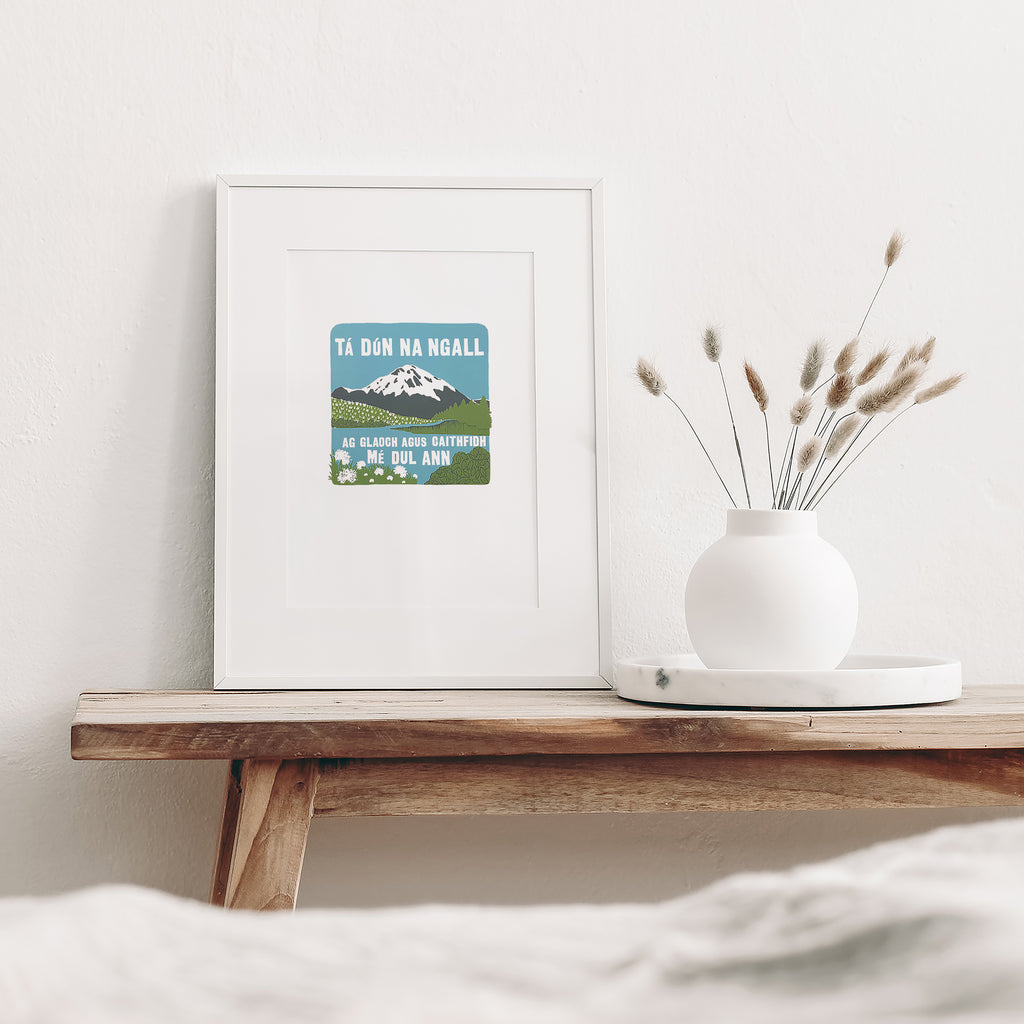 Irish Donegal screen print mounted in a white frame leaning against a white wall. It is sitting on a wood bench beside a white vase with dried flowers