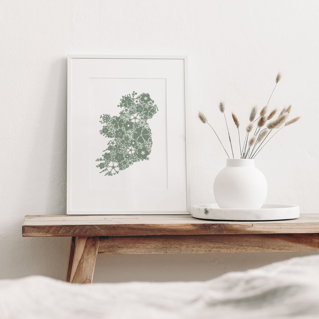 Green floral Ireland mounted in a white frame leaning against a white wall. It is sitting on a wood bench beside a white vase with dried flowers