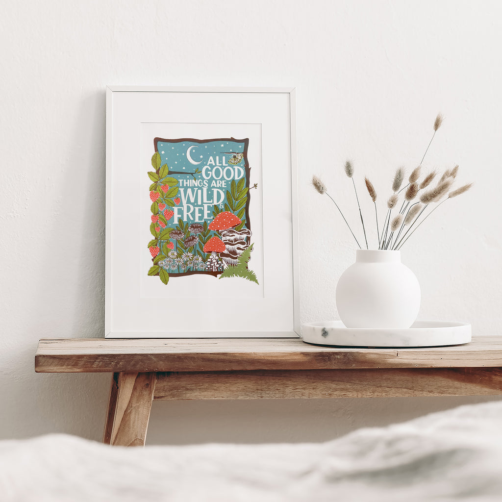 wild and free screen print mounted in a white frame leaning against the wall and sitting on a wood bench beside a white vase with dried flowers